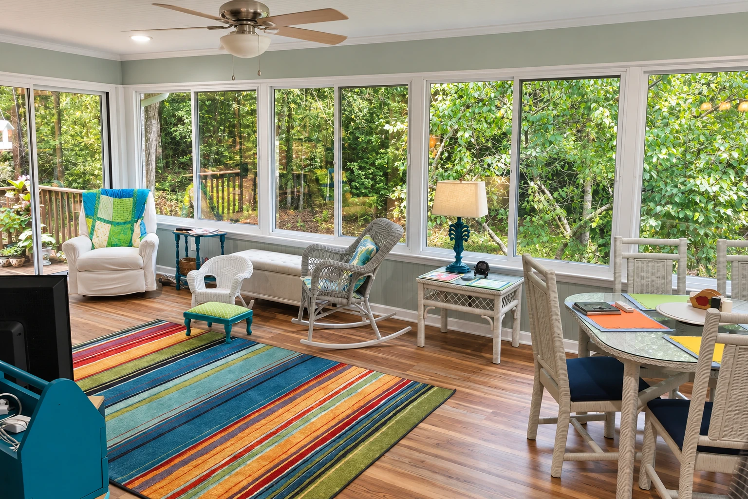 An enclosed patio room with white-framed windows, wicker seating, a dining table and a colorful striped rug overlooks leafy trees.