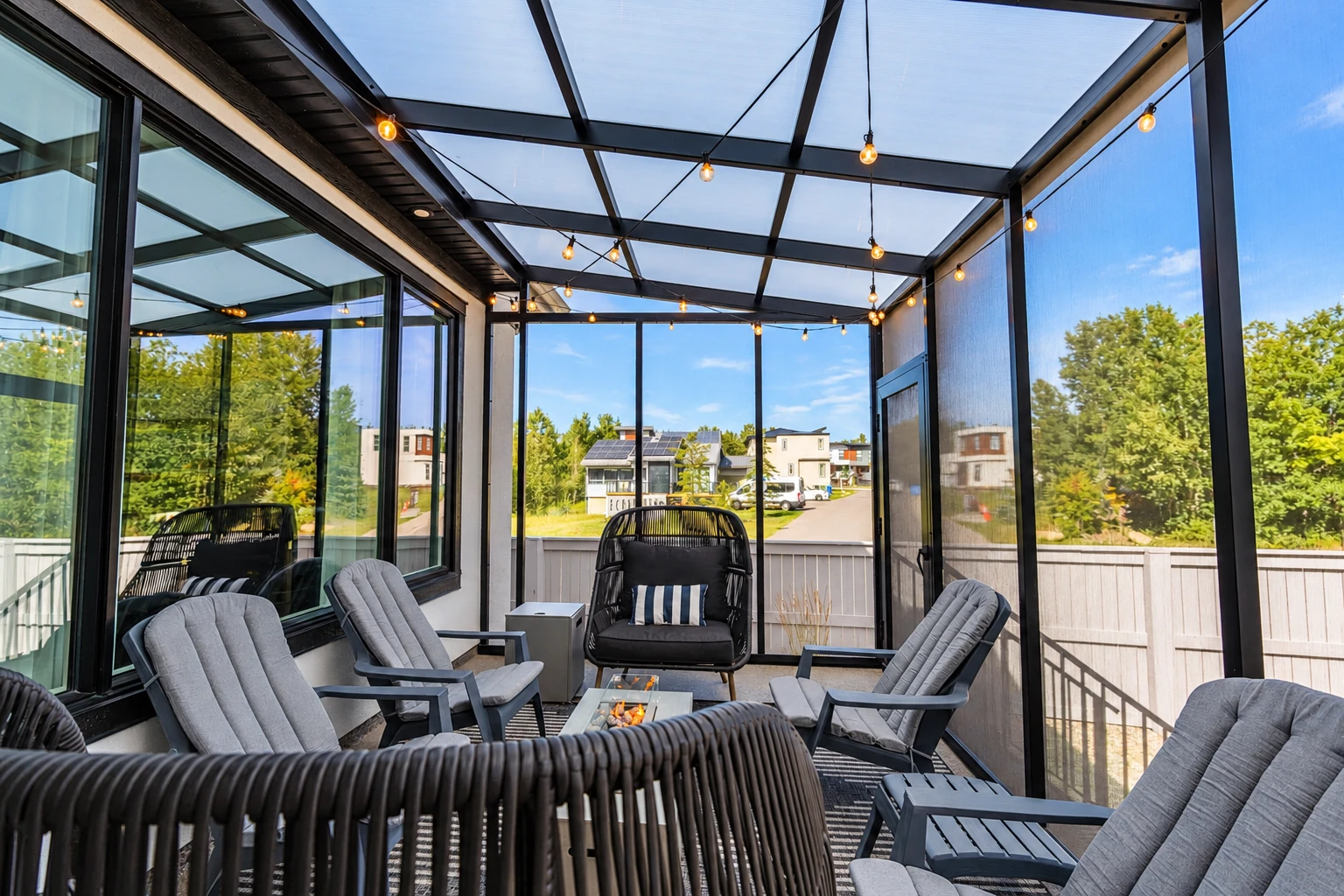 Enclosed patio with a clear roof, dark framing, string lights, and cushioned lounge chairs.