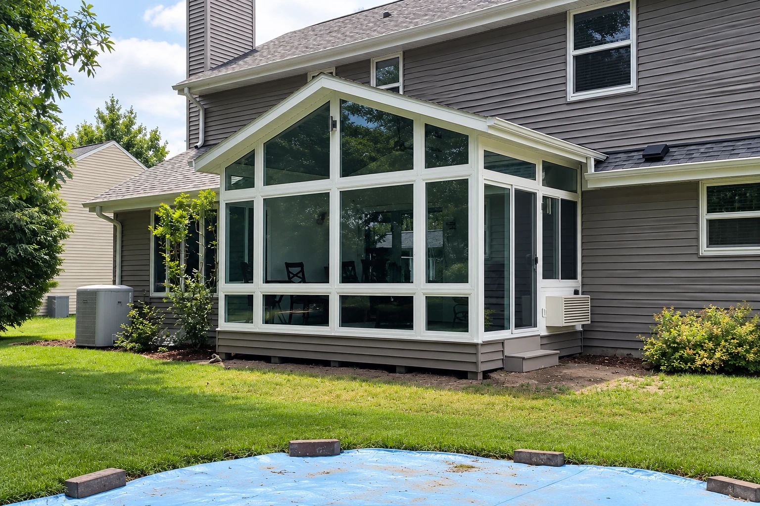 White-framed gable sunroom extends from a gray-sided house, with glass walls overlooking a lawn and trees.