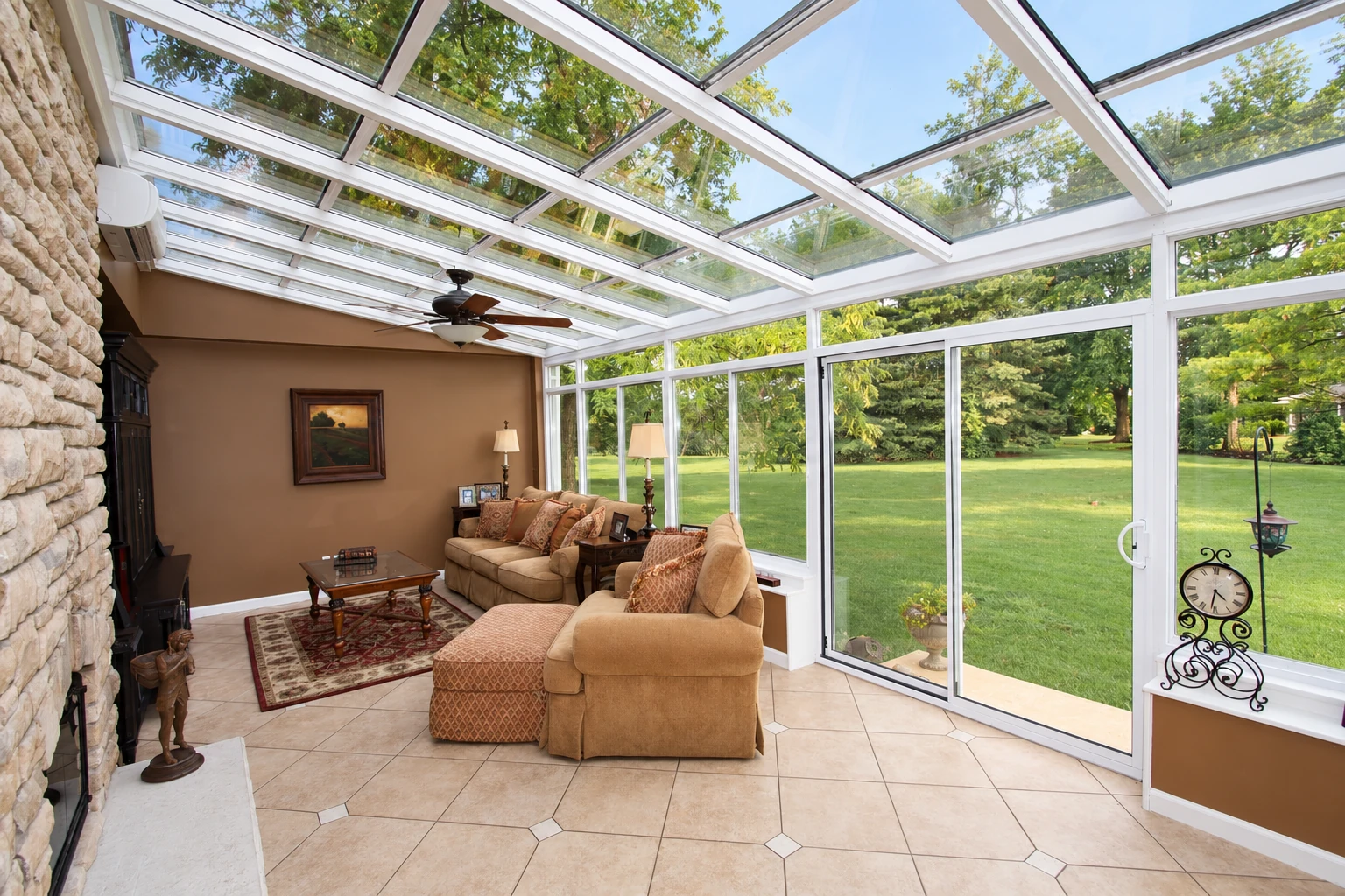 A furnished solarium with a glass roof and white-framed walls opens onto a green lawn and garden.