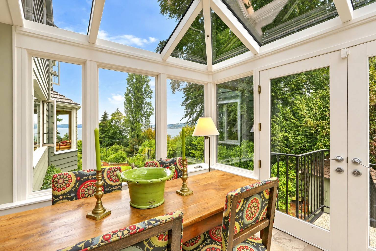 Bright sunroom with a glass roof, white frames, and a wooden dining table set with colorful chairs and a green centerpiece.