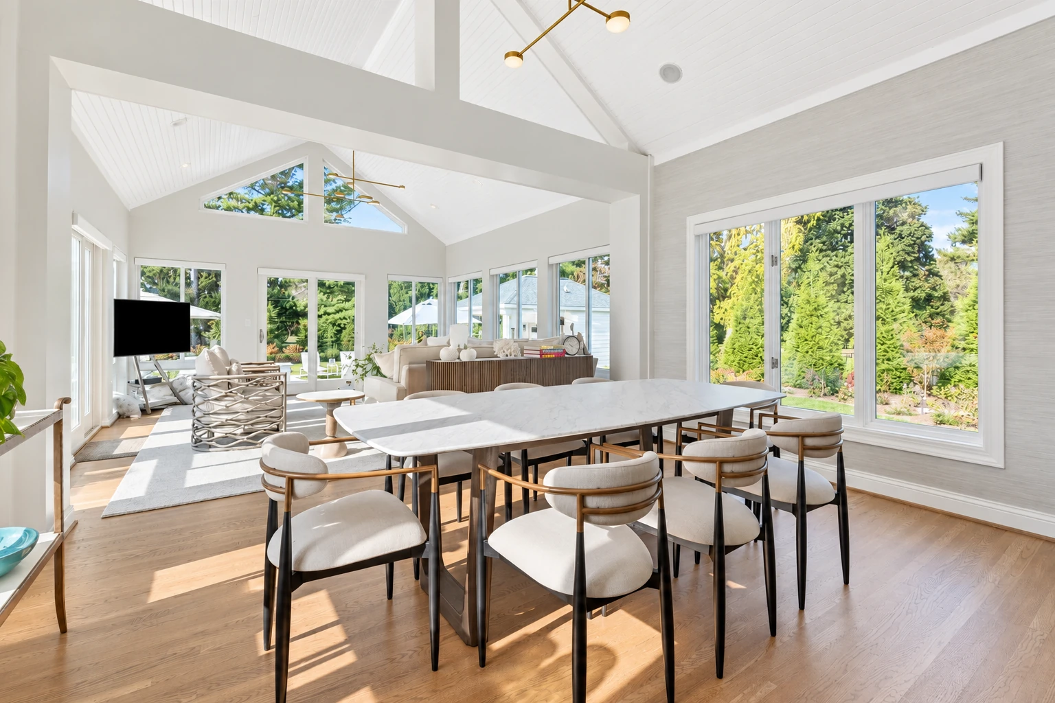 Bright sunroom with white walls, vaulted ceiling, expansive windows, and a dining table surrounded by chairs.